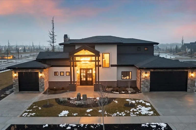 Modern house with stone and stucco exterior, illuminated entrance, two garage doors, landscaped front yard with patches of snow, and a sunset sky in the background.